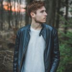 Young man in a black leather jacket standing in a forest during twilight, styled fashion portrait.